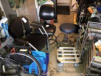 Photo showing two black arm chairs, black desk chair, blue rolling stool, metal rolling cart, and background items like a blue storage container.