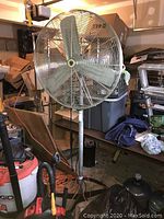 Full view of the 32 inch diameter industrial floor fan on adjustable height metal stand with tripod base in a cluttered storage space.
