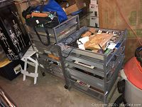 Two Elfa metal mesh storage units with drawers, one with wheels and one without, placed in a garage or storage area with various items on and inside the drawers.