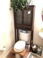 Front view of brown wooden over-the-toilet cabinet standing above toilet, showing two frosted glass doors and two drawers below.
