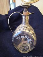 Front view of the 1940s dimple bottle decanter showing clear glass with 'DIMPLE' embossed and metal floral overlay and handle.