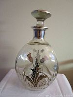 Side view of the clear glass dimpled bottle decanter showing painted thistle floral design and matching silver tone trimmed stopper.