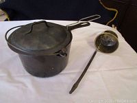 Cast iron pot with lid and brass water dipper laid side by side on a cloth surface.