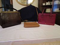 Three ladies leather handbags on white surface against dark background, showing all three handbags including dark brown crocodile texture bag, tan pouch, and reddish-pink handbag.