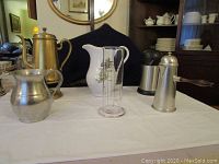 Various beverage servers and pitchers arranged on a white cloth-covered table in front of a dark backdrop and dining room furniture.