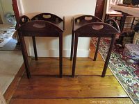 Pair of two dark wood hinged butler tray tables standing side by side on wood floor.