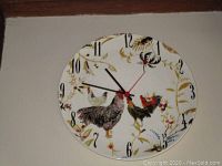 Round wall clock showing rooster and hen artwork on the face with black clock hands and red second hand on white background.