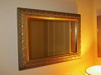 Wall mirror with gilt frame hung on wall, showing ornate carved leaf design and gold finish.