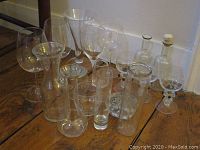 Group of various clear glassware items including wine glasses, carafes, vases, and decanters placed on wooden floor near wall baseboard.