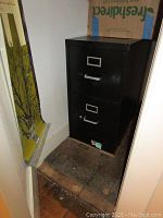 Black metal two drawer file cabinet placed on a tiled floor showing scuffs and wear.