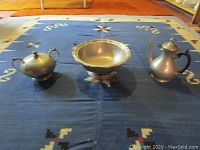 Overview of the three pewter pieces on a blue patterned rug including sugar bowl, pedestal bowl, and coffee pot.