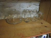 Photo showing the entire lot: glass cake dish with lid and three glass canisters with lids all sitting on a wooden shelf, against a stone wall background.