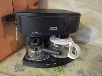 Front view of the KRUPS espresso/cappuccino maker showing the glass pot, steam wand, and machine controls.
