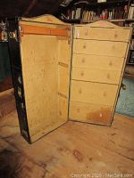 Vintage wardrobe trunk open showing hanging space with wooden bar and leather tab hangers on left, and five drawers with leather tab pulls on right.