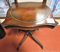 Front angled view of antique wooden end table showing leather top with gold embossing and carved wood apron and pedestal base.