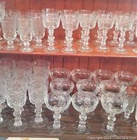 Shelf display of multiple rows of clear vintage glass stemware with etched patterns, including drinking and dessert glasses.