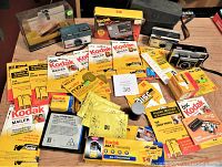 Full lot showing two vintage Kodak cameras, numerous unopened Kodak film discs in yellow mailers, film cartridge boxes, and a black Kodak camera case on a wood surface.