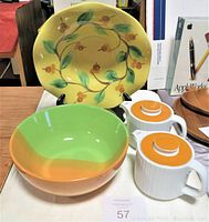 Top-side photo showing four ceramic kitchenware pieces: a large yellow bowl with floral design behind, a green and orange bowl in front, and two white and orange creamers with lids on the right side.