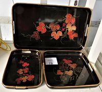 Three black lacquer trays with red and gold floral and vine patterns arranged on a floor for display. The trays vary in size and each has side handles.