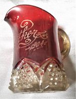 Photo of ruby red glass pitcher vase etched with 'Mother 1904'. Diamond cut clear base and clear handle visible.