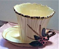 Side angle showing scalloped rim and floral handle attached to pale yellow planter with attached saucer plate.