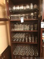 Full view of cabinet shelves displaying a large assortment of clear glass beverage glasses including etched and non-etched varieties with various shapes and sizes.