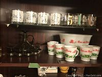 Shelf with six clear glasses with metallic gold and black numbered designs on the top shelf. Below is a white Tom and Jerry punch bowl and six matching white glasses with red and green scenes and text. To the left is a dark decanter with handle and stopper with six matching small glasses.