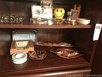 Shelved collection of assorted ashtrays made of ceramic, pottery, glass, and metal, plus bar accessories including a yellow smiley face container and small metal egg cup.