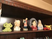 Seven hand-painted ceramic figurines including Royal Copley girl in middle with a small chip on left elbow, displayed on shelf.
