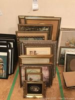 Stacks of various framed pictures and picture frames leaning against a wall, showing size variety and frame styles