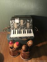 Camillo Jr. accordion on top of a grayish blue carrying case, four maracas and small bongos in front on wooden floor
