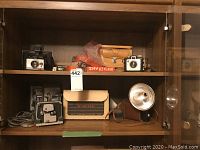 Wooden cabinet shelf holding vintage cameras and related items: Polaroid Colour Pack camera, Brownie Holiday Flash camera with case, Primo 8mm film editor, Brownie Hawkeye camera with broken case clasp.