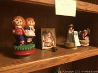 Wide angle of six different music boxes arranged on wooden shelf including two figurine children, framed girl, woman in bonnet, man with hat, and seated figure.