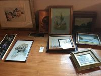 Wide view of 9 framed artworks and prints on a wooden floor, including portrait, landscape, and still life subjects.