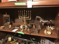 Shelf display of various bronze-coloured metal items including menorah, door bells, horse figurine, and small decorative pieces.