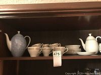 Shelf with a porcelain blue and white geometric teapot, several white cups with gold fern decoration, and a stack of white plates with gold detail.