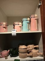 Shelf displaying four ceramic air-tight canisters in pastel pink and green with wooden scoops on the left side of shelf below are pink and green ceramic soup bowls with handles and lids.