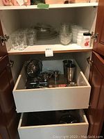 Open cabinet showing clear glasses, white bowls, Pyrex pie plates with lids on top shelf, stainless steel serving ware in drawer below