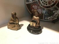 Two Asian-style figurines: one a bronze-colored Buddha figure seated, the other gold-colored figurine seated with a box, displayed on carved wooden stand.