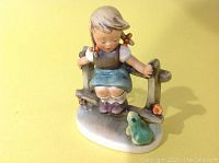Front view of the Hummel girl figurine sitting on a fence looking at a green frog at her feet.