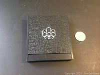 Closed black textured box with Montreal Olympics emblem showing the original packaging of the commemorative coin.