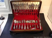Opened hardwood presentation chest with red lining showing full flatware set arranged in slots.