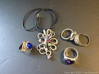 Full view showing four sterling silver rings and one abstract pendant on a black cord with colored stones.
