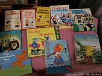 Photo of 10 children's books including Charlie Brown titles, Popeye, Wizard of Oz, and others on a pink surface.