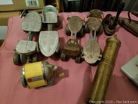 Photo shows seven pairs of vintage metal roller skate attachments and a brass cylindrical container on a pink surface with other vintage tools and irons nearby.