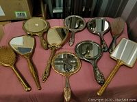 Vintage handheld mirrors and hair brushes laid out on a pink cloth, showing assorted styles, shapes, and metallic finishes.