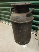 Front view of a heavy vintage iron milk can, showing cylindrical body, two handles, and wide mouth opening.