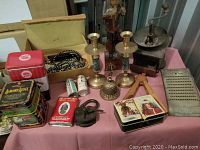Photo shows an assortment of vintage household and kitchen items on a pink cloth-covered table, including colorful tins, a cast iron coffee grinder, brass candlestick holders, a hand bell, a wrought iron lock, an iron, a large metal grater, and a wooden figurine.