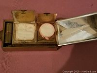 Open vintage wristlet makeup kit showing compartments with powder compact, powder puff, lipstick container, and visible interior mirror
