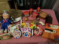 Wide shot showing full assortment of items including doll, canteen, toy revolver, noise makers, clocks, pipe, and more arranged on pink cloth surface.